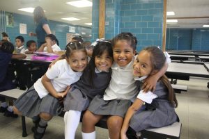 Four young girls hug and smile for the camera as they sit in their classroom.